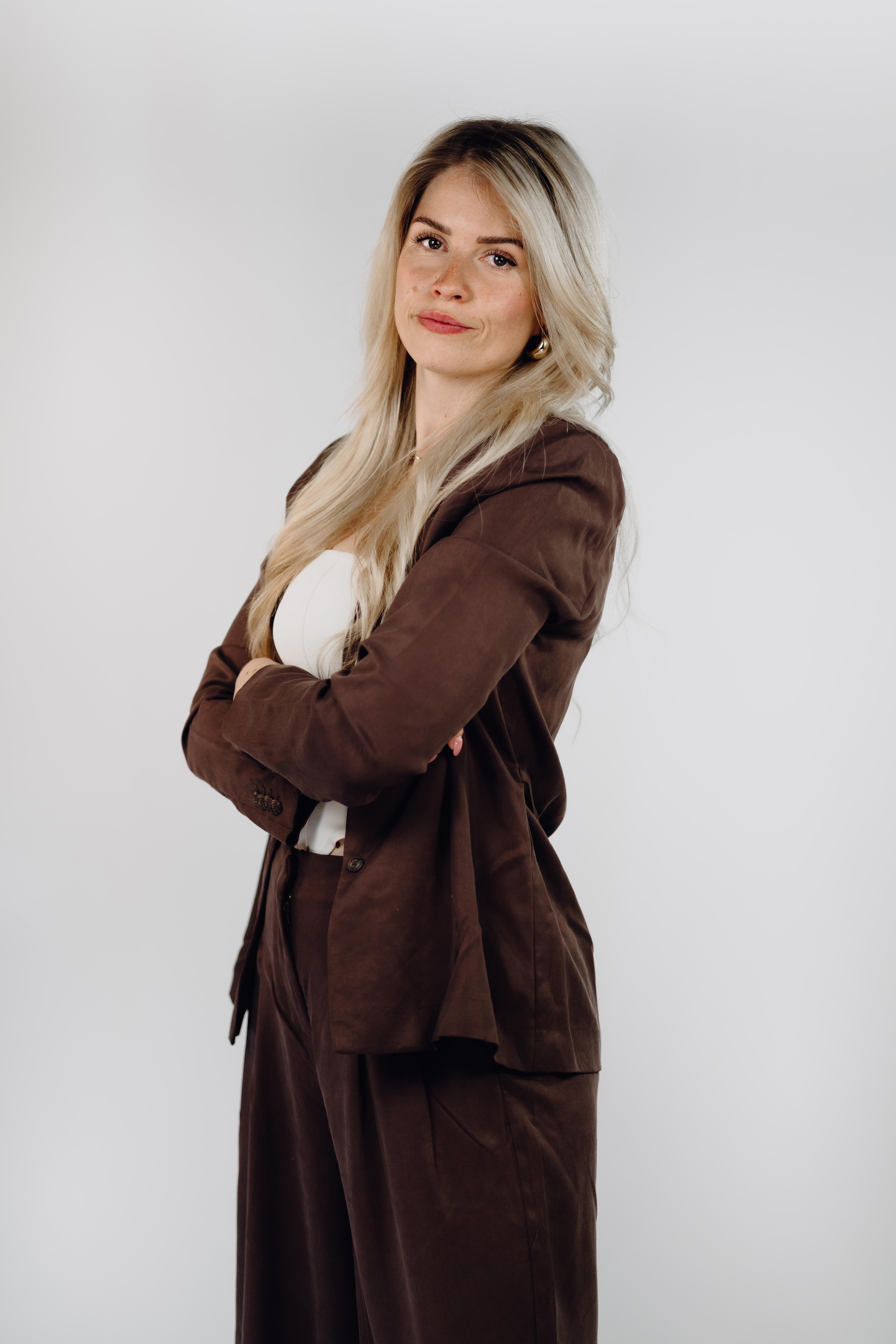 Professionelles Porträtfoto einer Frau mit langen blonden Haaren, seitlich stehend mit verschränkten Armen, in einem braunen Blazer mit passender weiter Hose.