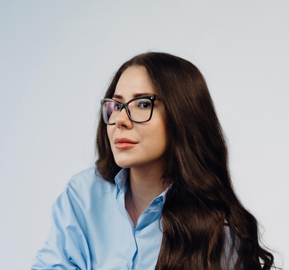 Professional headshot of a woman with long dark wavy hair and glasses, wearing a light blue blouse and dark jeans, arms crossed, smiling slightly at the camera.