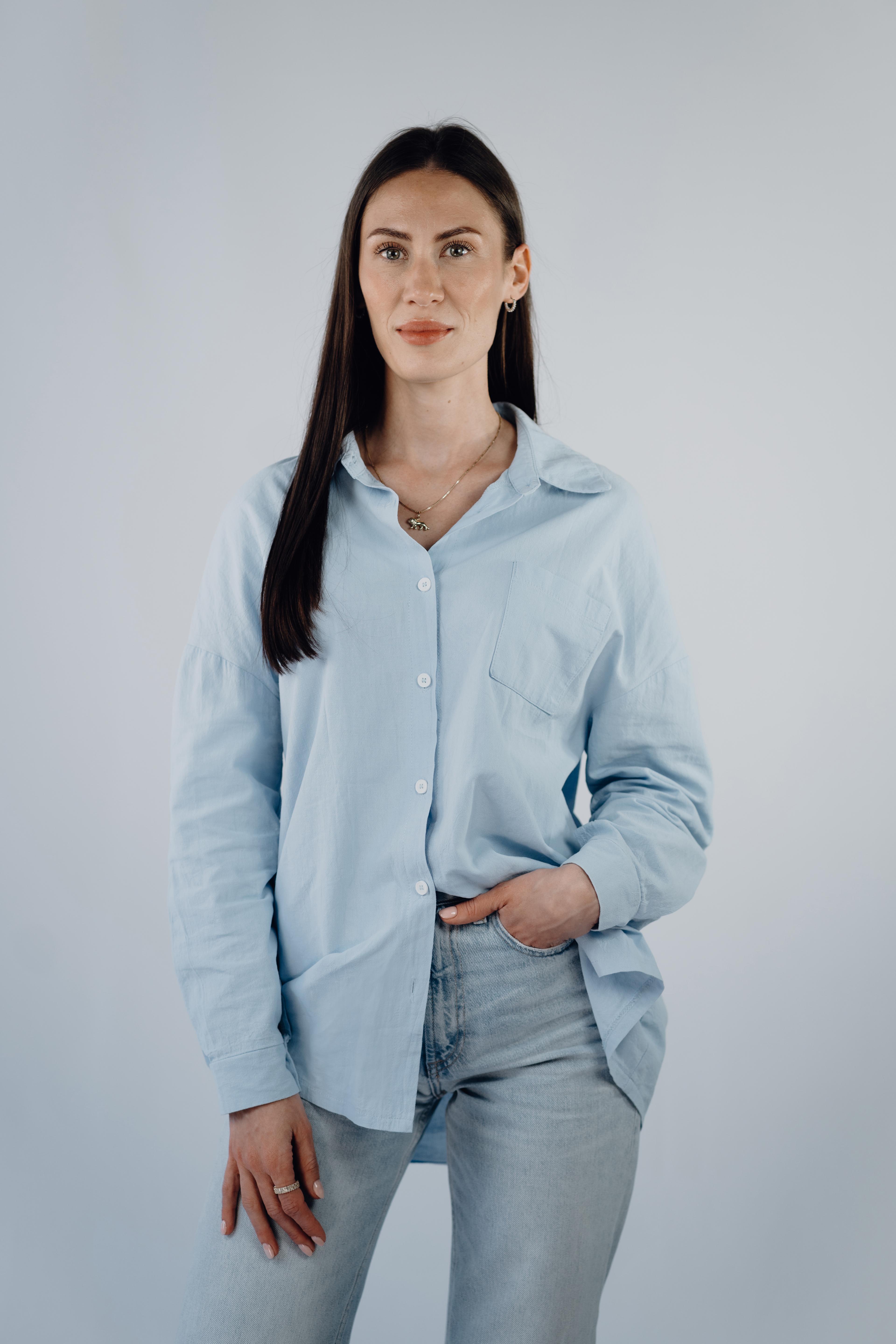 Professionelles Porträtfoto einer Frau mit langen glatten dunklen Haaren, in einem hellblauen Hemd und Jeans, eine Hand in der Tasche, freundlich in die Kamera blickend.