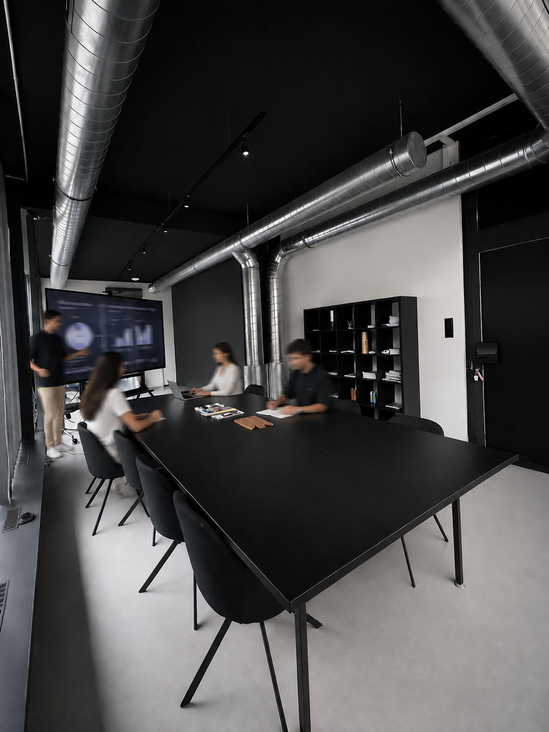 Moderner Besprechungsraum mit schwarzem Konferenztisch und Stühlen, freiliegenden silbernen Lüftungsrohren an einer schwarzen Decke und vier Kollegen in einer Präsentation, einer davon steht vor einem an der Wand montierten Bildschirm, auf dem Diagramme angezeigt werden, die restlichen Kollegen sitzen beim Konferenztisch.