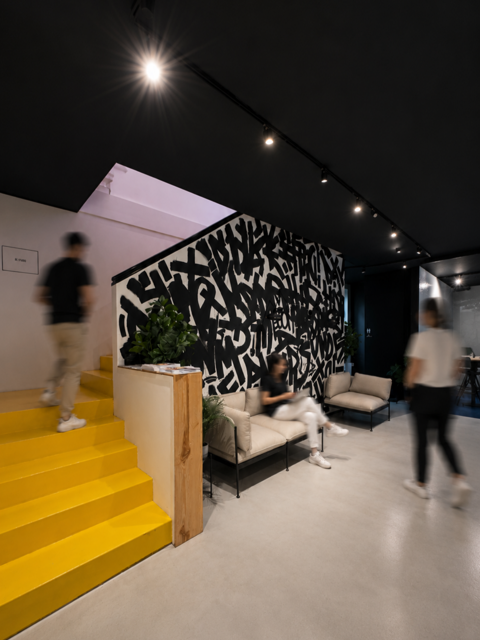 Bürolobby mit leuchtend gelber Treppe und einer schwarz-weißen Akzentwand im Graffiti-Stil, eine Person sitzt auf einem beigen Sofa und zwei andere gehen in Bewegungsunschärfe vorbei, alles unter warmem Scheinwerferlicht.