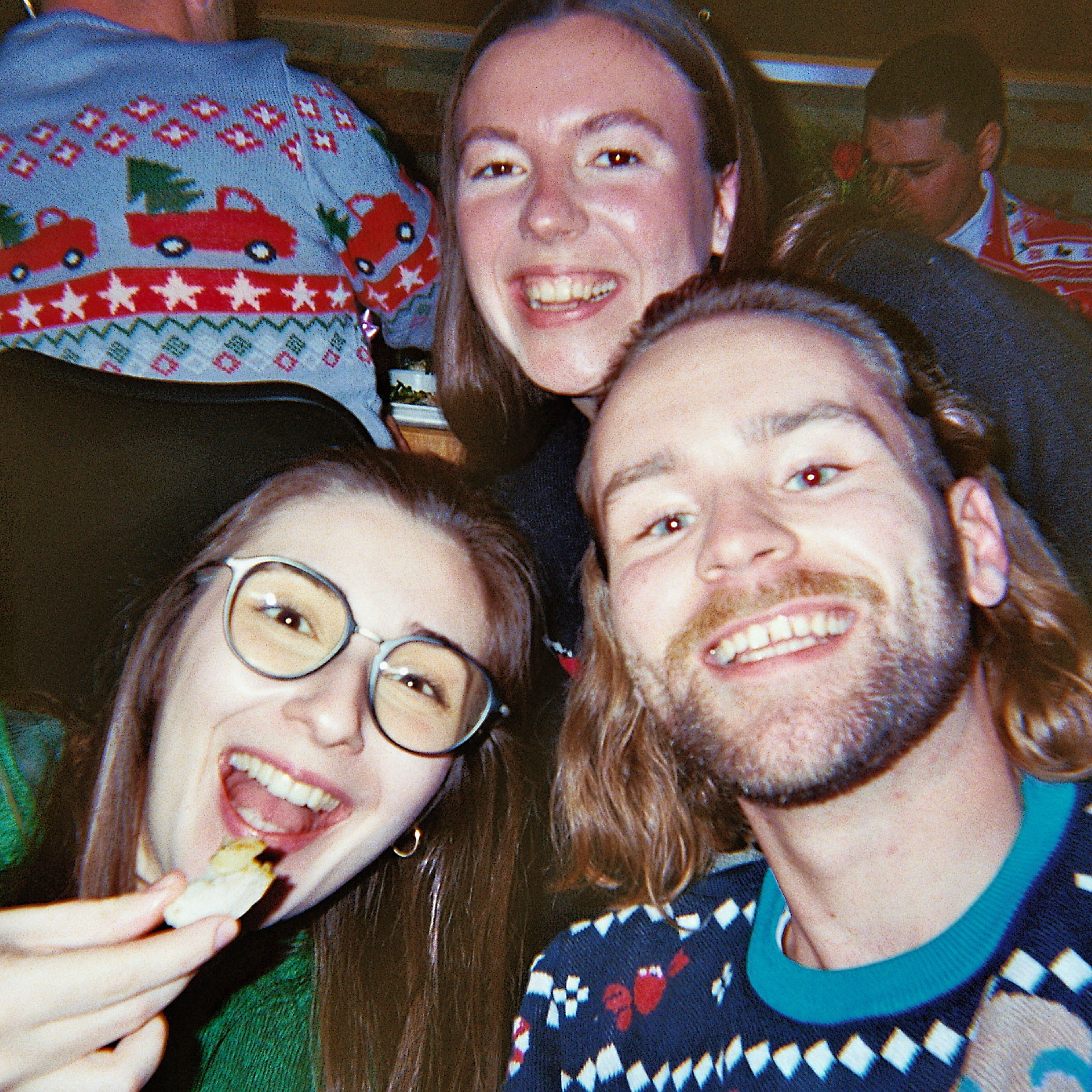 Ein Nahaufnahme-Selfie von drei Freunden, die auf einer festlichen Party lächeln; zwei von ihnen tragen Pullover mit Weihnachtsmotiven, einer isst einen Snack, im Hintergrund ist eine verschwommene Menschenmenge zu sehen.