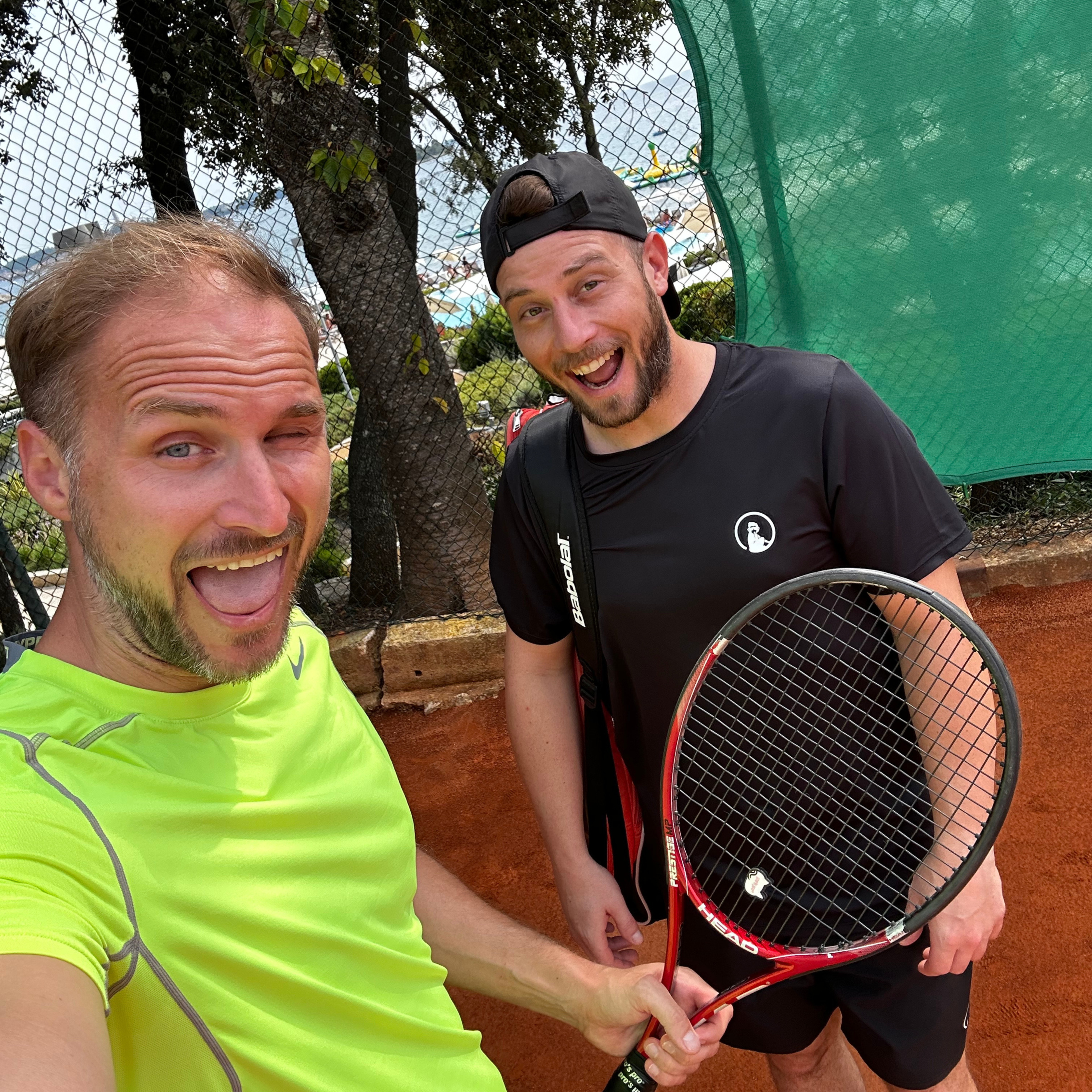 Zwei Männer machen ein Selfie auf einem roten Sandtennisplatz, einer in einem leuchtend gelbgrünen Hemd und der andere in Schwarz mit einem Tennisschläger in der Hand, beide lächeln vor dem Hintergrund von Bäumen und einem grünen Zaun.